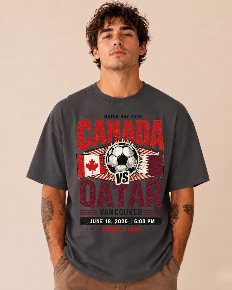 May include: Dark gray t-shirt with a distressed graphic featuring "CANADA vs QATAR" with a soccer ball, flags, and event details. The text includes "VANCOUVER JUNE 18, 2026 | 6:00 PM".