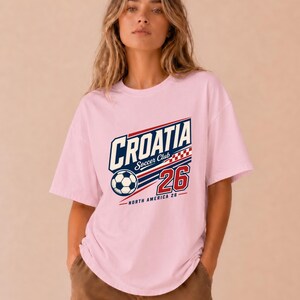 K&ouml;nnte beinhalten: Ein hellrosa T-Shirt mit kurzen &Auml;rmeln und einem Grafikdesign, das den Text CROATIA Soccer Club, die Zahl 26, einen Fu&szlig;ball und ein rot-wei&szlig;es Karomuster zeigt. Der Text NORTH AMERICA 26 ist in kleineren Buchstaben unter der Grafik gedruckt.