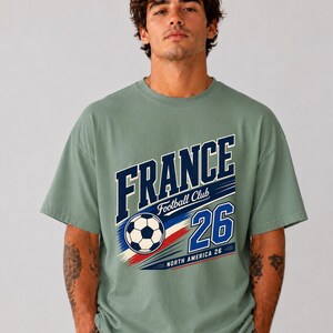 May include: A sage green short-sleeve t-shirt featuring a graphic print with the text FRANCE Football Club 26 North America 26 and a football illustration. The design includes blue, white, and red brushstroke accents across the chest.