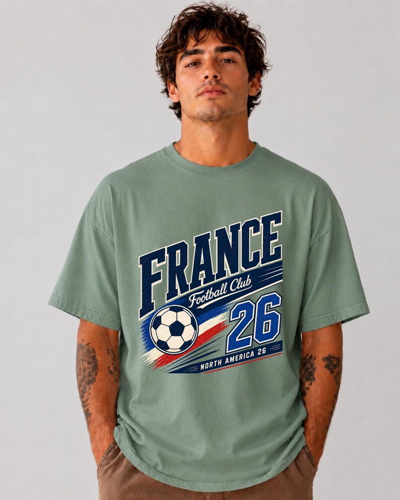 May include: A sage green short-sleeve t-shirt featuring a graphic print with the text FRANCE Football Club 26 North America 26 and a football illustration. The design includes blue, white, and red brushstroke accents across the chest.
