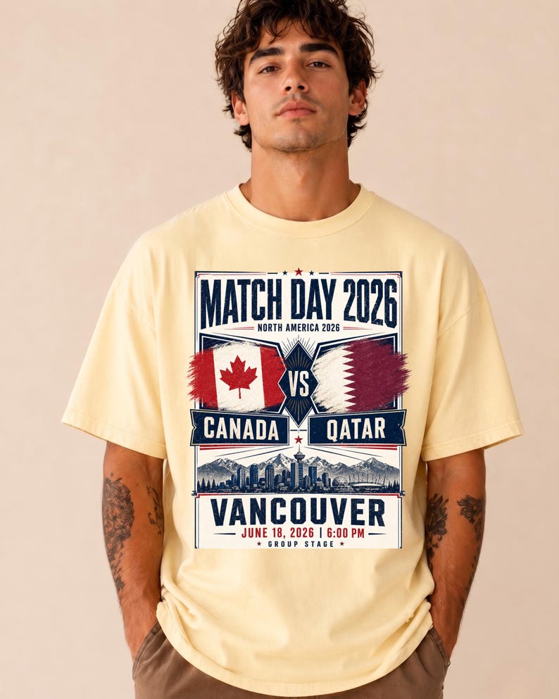 May include: A light yellow t-shirt featuring a graphic for the 2026 Match Day in North America. The design includes the flags of Canada and Qatar, the city of Vancouver, and the date and time of the match.
