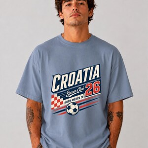 以下が含まれることがあります： CROATIA Soccer Club 26 NORTH AMERICA 26と書かれたグラフィックプリントが施された、ダスティブルーの半袖Tシャツ。デザインには、赤と白の市松模様の正方形、サッカーボール、赤と青の動きを表す線が含まれています。