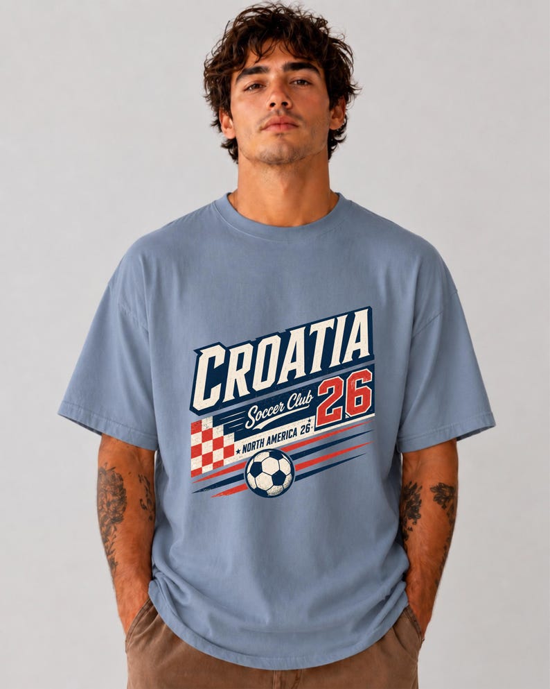 以下が含まれることがあります： CROATIA Soccer Club 26 NORTH AMERICA 26と書かれたグラフィックプリントが施された、ダスティブルーの半袖Tシャツ。デザインには、赤と白の市松模様の正方形、サッカーボール、赤と青の動きを表す線が含まれています。