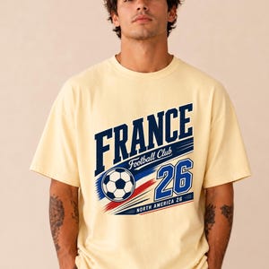 May include: A cream-coloured short-sleeve t-shirt featuring a graphic print with the text FRANCE Football Club and the number 26. The design includes a football illustration, blue and red brushstroke accents, and the text NORTH AMERICA 26.