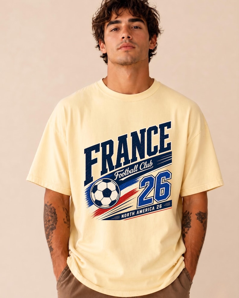 May include: A cream-coloured short-sleeve t-shirt featuring a graphic print with the text FRANCE Football Club and the number 26. The design includes a football illustration, blue and red brushstroke accents, and the text NORTH AMERICA 26.