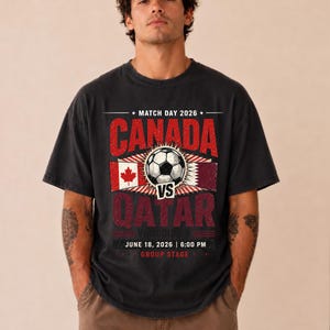 May include: Black t-shirt with a soccer-themed graphic. The design features the text "CANADA" and "QATAR" with a soccer ball in the center. Additional text includes "MATCH DAY 2026", "VANCOUVER", "JUNE 18, 2026 | 6:00 PM", and "GROUP STAGE".