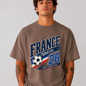 May include: A brown short-sleeve t-shirt featuring a graphic print with the text FRANCE, Football Club, 26, and NORTH AMERICA 26. The design includes a football and blue, white, and red brushstroke accents across the centre of the chest.