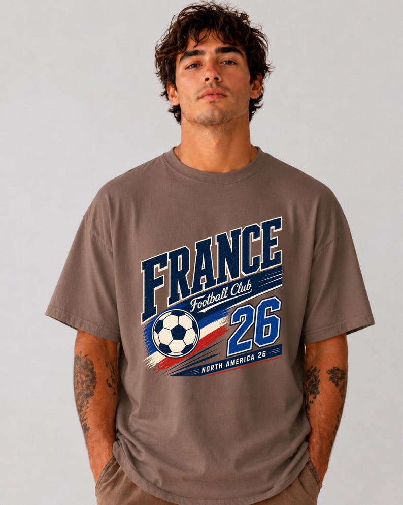 May include: A brown short-sleeve t-shirt featuring a graphic print with the text FRANCE, Football Club, 26, and NORTH AMERICA 26. The design includes a football and blue, white, and red brushstroke accents across the centre of the chest.