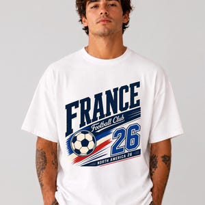 May include: A white short-sleeve t-shirt featuring a graphic print with the text FRANCE, Football Club, and 26. The design includes a football, blue and red brush strokes, and the text NORTH AMERICA 26 in a bold, athletic font style.