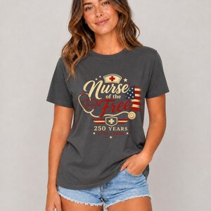May include: Dark gray t-shirt with the text "Nurse of the Free" in a vintage design, featuring a stethoscope, cross, and American flag. The shirt also includes the text "250 Years 1776-2026".
