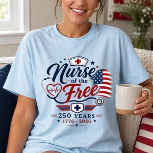 May include: Light blue t-shirt with the text "Nurse of the Free" in navy blue script, a red heart with a heartbeat, and an American flag design. The shirt also features a nurse's cap and the text "250 Years 1776-2026."