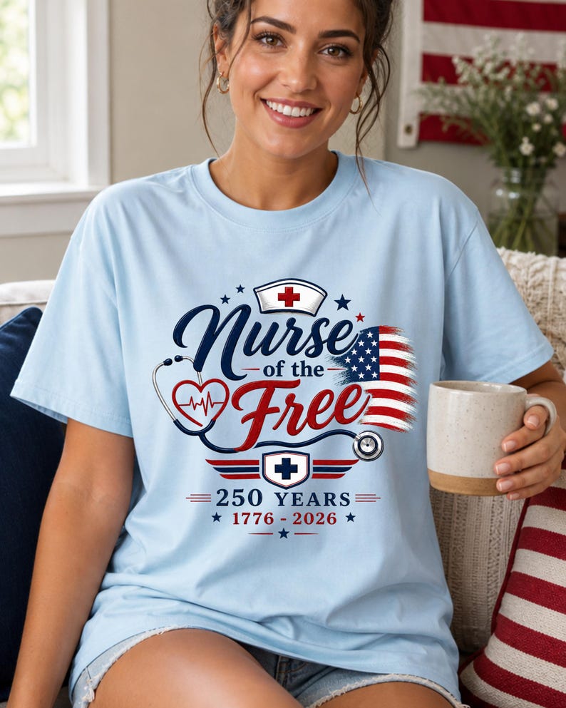 May include: Light blue t-shirt with the text "Nurse of the Free" in navy blue script, a red heart with a heartbeat, and an American flag design. The shirt also features a nurse's cap and the text "250 Years 1776-2026."