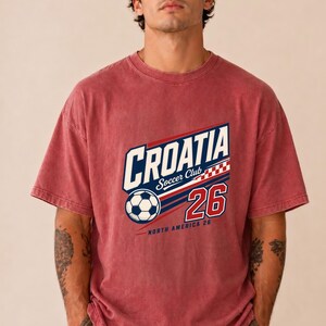 K&ouml;nnte beinhalten: Ein verwaschenes rotes Kurzarm-T-Shirt mit einem Grafikdruck, der CROATIA Soccer Club zeigt, zusammen mit einem Fu&szlig;ball, einem rot-wei&szlig;en Karomuster, der Zahl 26 und dem Text NORTH AMERICA 26 in den Farben Blau, Wei&szlig; und Rot.
