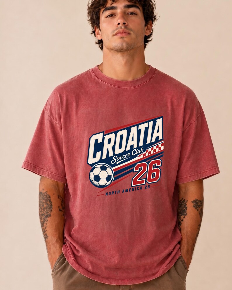 K&ouml;nnte beinhalten: Ein verwaschenes rotes Kurzarm-T-Shirt mit einem Grafikdruck, der CROATIA Soccer Club zeigt, zusammen mit einem Fu&szlig;ball, einem rot-wei&szlig;en Karomuster, der Zahl 26 und dem Text NORTH AMERICA 26 in den Farben Blau, Wei&szlig; und Rot.