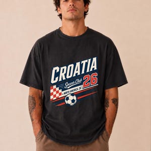 以下が含まれることがあります： CROATIA Soccer Club 26 NORTH AMERICA 26と書かれたグラフィックプリントが施されたチャコールグレーの半袖Tシャツ。デザインには、赤と白の市松模様の正方形、サッカーボール、赤と青の水平ストライプが含まれています。