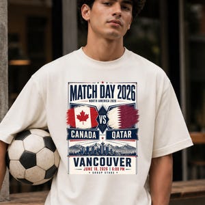May include: Cream-colored t-shirt featuring a soccer ball and a graphic for the 2026 Match Day in Vancouver. The design includes the flags of Canada and Qatar, with text indicating the date and time of the match.