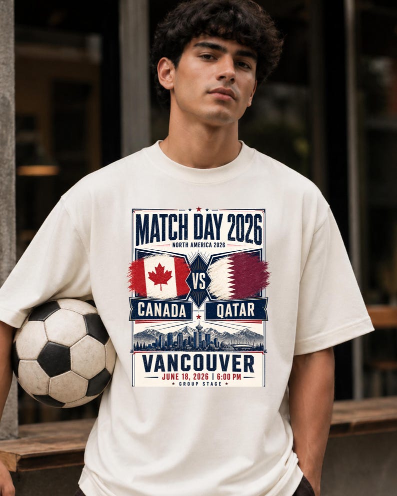 May include: Cream-colored t-shirt featuring a soccer ball and a graphic for the 2026 Match Day in Vancouver. The design includes the flags of Canada and Qatar, with text indicating the date and time of the match.