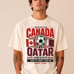 May include: Cream-colored t-shirt featuring a soccer ball graphic with the text "CANADA vs QATAR" in red and maroon. The shirt also includes the date "JUNE 18, 2026" and the location "VANCOUVER".