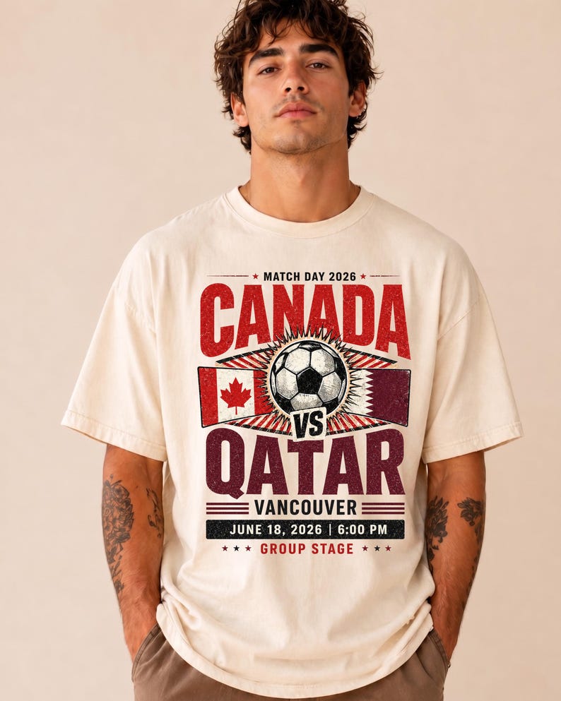 May include: Cream-colored t-shirt featuring a soccer ball graphic with the text "CANADA vs QATAR" in red and maroon. The shirt also includes the date "JUNE 18, 2026" and the location "VANCOUVER".