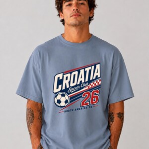K&ouml;nnte beinhalten: Ein staubblaues Kurzarm-T-Shirt mit einem Grafikdruck, der CROATIA Soccer Club, einen Fu&szlig;ball, ein rot-wei&szlig;es Karomuster, die Zahl 26 und den Text NORTH AMERICA 26 in roter, wei&szlig;er und dunkelblauer Tinte zeigt.