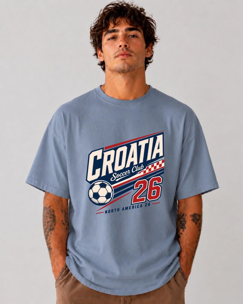 K&ouml;nnte beinhalten: Ein staubblaues Kurzarm-T-Shirt mit einem Grafikdruck, der CROATIA Soccer Club, einen Fu&szlig;ball, ein rot-wei&szlig;es Karomuster, die Zahl 26 und den Text NORTH AMERICA 26 in roter, wei&szlig;er und dunkelblauer Tinte zeigt.