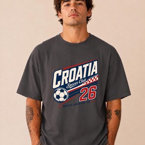 K&ouml;nnte beinhalten: Ein anthrazitfarbenes Kurzarm-T-Shirt mit einem Grafikdruck, der "CROATIA Soccer Club" in wei&szlig;en und blauen Buchstaben zeigt, dazu ein Fu&szlig;ball, ein rot-wei&szlig;es Karomuster, die Zahl "26" in Rot sowie der Schriftzug "NORTH AMERICA 26" in kleiner Schrift.