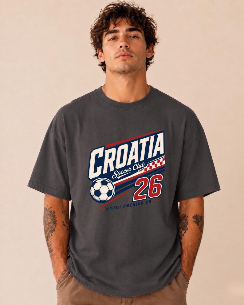 K&ouml;nnte beinhalten: Ein anthrazitfarbenes Kurzarm-T-Shirt mit einem Grafikdruck, der "CROATIA Soccer Club" in wei&szlig;en und blauen Buchstaben zeigt, dazu ein Fu&szlig;ball, ein rot-wei&szlig;es Karomuster, die Zahl "26" in Rot sowie der Schriftzug "NORTH AMERICA 26" in kleiner Schrift.