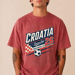 以下が含まれることがあります： CROATIA Soccer Club 26 NORTH AMERICA 26と書かれたグラフィックプリントが施された、色あせた赤の半袖Tシャツ。デザインには、赤と白の市松模様、サッカーボール、青と赤の水平な動きの線が含まれています。