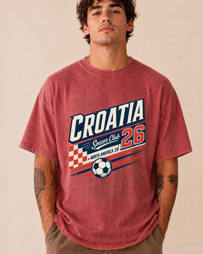 以下が含まれることがあります： CROATIA Soccer Club 26 NORTH AMERICA 26と書かれたグラフィックプリントが施された、色あせた赤の半袖Tシャツ。デザインには、赤と白の市松模様、サッカーボール、青と赤の水平な動きの線が含まれています。