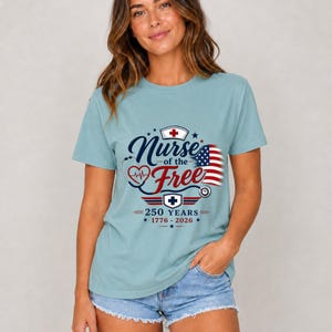 May include: A light blue t-shirt with the text "Nurse of the Free" in navy blue script, featuring a red heart, stethoscope, and American flag design. The shirt also includes the text "250 YEARS 1776-2026".