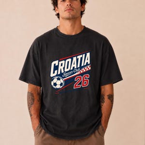 K&ouml;nnte beinhalten: Ein anthrazitfarbenes Kurzarm-T-Shirt mit einem Grafikdruck, der CROATIA Soccer Club mit einem Fu&szlig;ball-Symbol, einem rot-wei&szlig;en Karomuster und der Zahl 26 in Rot mit wei&szlig;er Umrandung auf der Vorderseite zeigt.