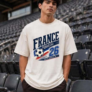 May include: An off-white short-sleeve t-shirt featuring a graphic print that reads FRANCE Football Club 26 North America 26, accompanied by a football illustration and red, white, and blue motion lines across the chest area.