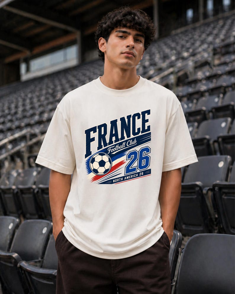 May include: An off-white short-sleeve t-shirt featuring a graphic print that reads FRANCE Football Club 26 North America 26, accompanied by a football illustration and red, white, and blue motion lines across the chest area.
