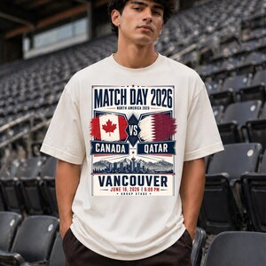 May include: A cream-colored t-shirt with a graphic promoting a soccer match. The design includes the text "MATCH DAY 2026" and "CANADA VS QATAR" with flags. The location and date are also displayed: "VANCOUVER JUNE 18, 2026 | 6:00 PM".