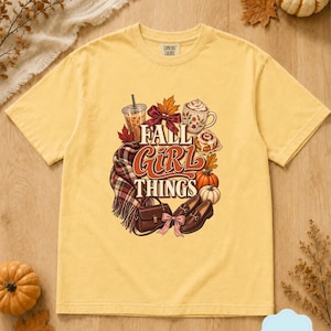 May include: A pale yellow short-sleeve t-shirt featuring a graphic print with the text "FALL GIRL THINGS" surrounded by illustrations of an iced coffee, a mug, a cinnamon roll, pumpkins, a plaid scarf, a handbag, and brown loafers.