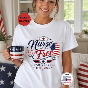 May include: White t-shirt with a patriotic design featuring the text "Nurse of the Free" in navy blue and red, an American flag, and a stethoscope graphic. The shirt also includes the text "250 YEARS 1776-2026".