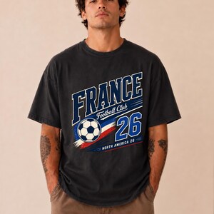 May include: A charcoal grey short-sleeve t-shirt featuring a graphic print with the text FRANCE, Football Club, and 26. The design includes a football, red, white, and blue brush strokes, and the text NORTH AMERICA 26 in a bold font.