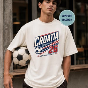 K&ouml;nnte beinhalten: Ein cremefarbenes Kurzarm-T-Shirt mit einer Grafik, die den Text CROATIA Soccer Club, die Zahl 26, einen Fu&szlig;ball und ein rot-wei&szlig;es Karomuster zeigt. Das Design enth&auml;lt den Text NORTH AMERICA 26 sowie ein Comfort Colors-Logo.