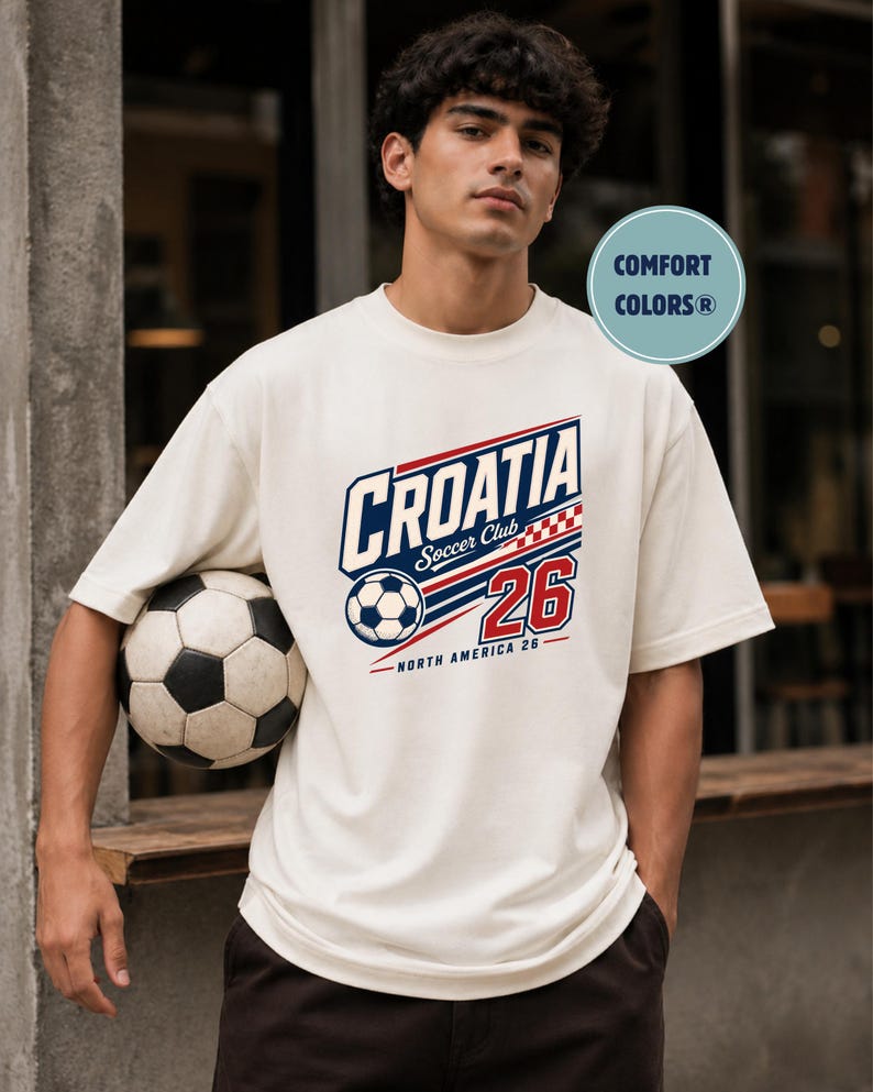 K&ouml;nnte beinhalten: Ein cremefarbenes Kurzarm-T-Shirt mit einer Grafik, die den Text CROATIA Soccer Club, die Zahl 26, einen Fu&szlig;ball und ein rot-wei&szlig;es Karomuster zeigt. Das Design enth&auml;lt den Text NORTH AMERICA 26 sowie ein Comfort Colors-Logo.