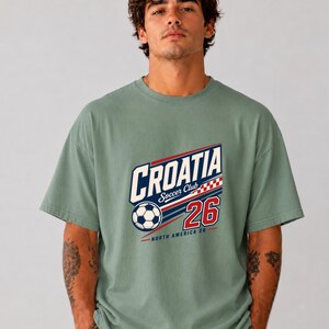 K&ouml;nnte beinhalten: Ein kurz&auml;rmeliges T-Shirt in Salbeigr&uuml;n mit einem Grafikdruck, der "CROATIA Soccer Club" in blauen und wei&szlig;en Buchstaben, einen Fu&szlig;ball, ein rot-wei&szlig;es Karomuster, die Zahl "26" sowie "NORTH AMERICA 26" in roter Schrift zeigt.