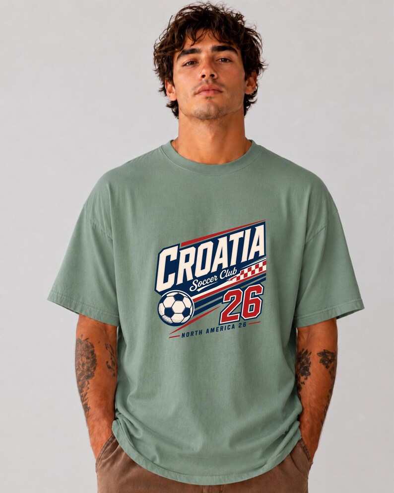 K&ouml;nnte beinhalten: Ein kurz&auml;rmeliges T-Shirt in Salbeigr&uuml;n mit einem Grafikdruck, der "CROATIA Soccer Club" in blauen und wei&szlig;en Buchstaben, einen Fu&szlig;ball, ein rot-wei&szlig;es Karomuster, die Zahl "26" sowie "NORTH AMERICA 26" in roter Schrift zeigt.