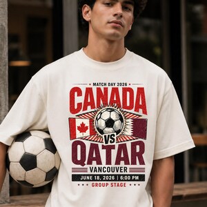May include: Cream-colored t-shirt featuring a soccer ball and the text "CANADA VS QATAR" in red and burgundy. The shirt includes the date "JUNE 18, 2026" and the location "VANCOUVER". A soccer ball is held in the arm.