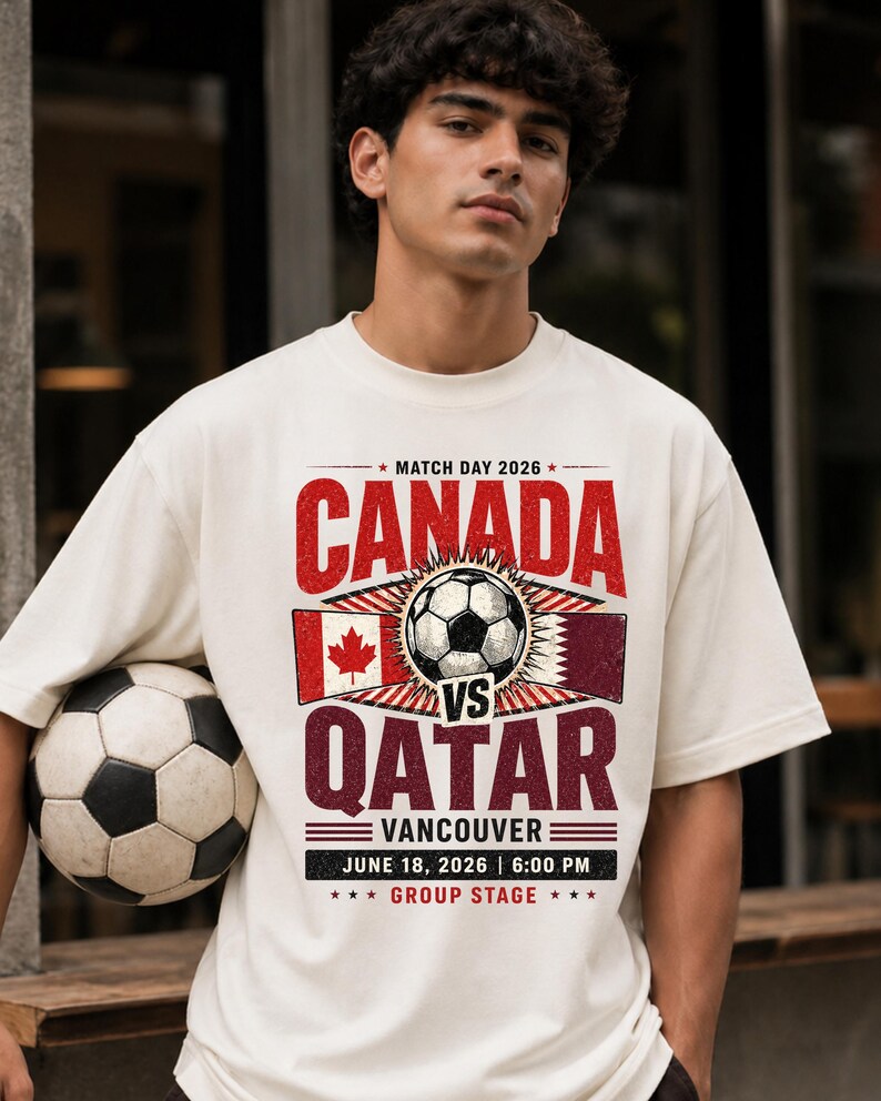 May include: Cream-colored t-shirt featuring a soccer ball and the text "CANADA VS QATAR" in red and burgundy. The shirt includes the date "JUNE 18, 2026" and the location "VANCOUVER". A soccer ball is held in the arm.