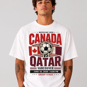 May include: White t-shirt featuring a soccer match graphic. The design includes the text "CANADA vs QATAR" with flags and a soccer ball. The text also includes "VANCOUVER", the date "JUNE 18, 2026", and "6:00 PM".