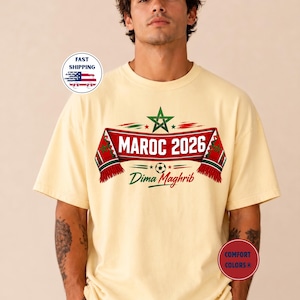 May include: Cream-colored t-shirt featuring a design with the text "MAROC 2026" in red and white, with a green star and the words "Dima Maghrib". The design includes a soccer ball graphic. The shirt has short sleeves.