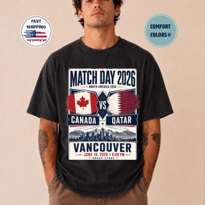 May include: Black t-shirt with a graphic for the 2026 Match Day between Canada and Qatar in Vancouver. The design includes the flags of Canada and Qatar, the date, and the time of the match.