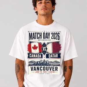 May include: White t-shirt with a graphic promoting a soccer match between Canada and Qatar in Vancouver. The design includes the text "Match Day 2026," flags, and the date June 18, 2026, at 6:00 PM. The shirt is worn by a person.