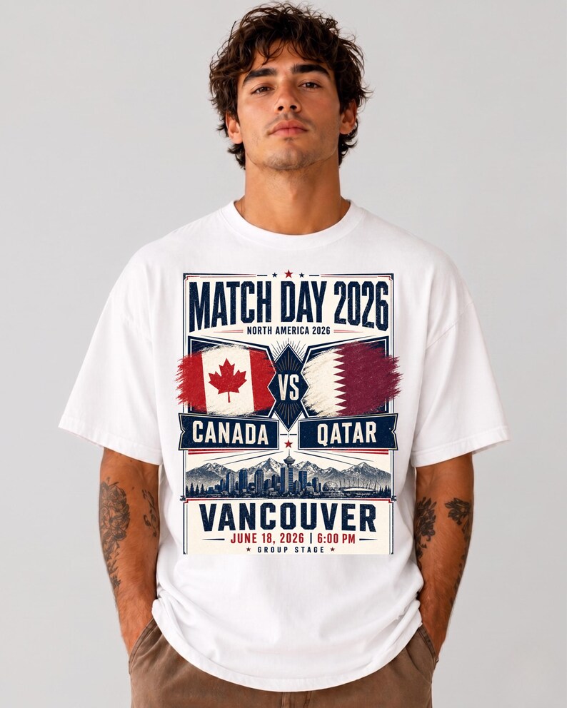 May include: White t-shirt with a graphic promoting a soccer match between Canada and Qatar in Vancouver. The design includes the text "Match Day 2026," flags, and the date June 18, 2026, at 6:00 PM. The shirt is worn by a person.