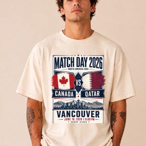May include: Cream-colored t-shirt featuring a graphic for a 2026 soccer match between Canada and Qatar in Vancouver. The design includes flags, city skyline, and the text "Match Day 2026". The date and time of the match are also displayed.