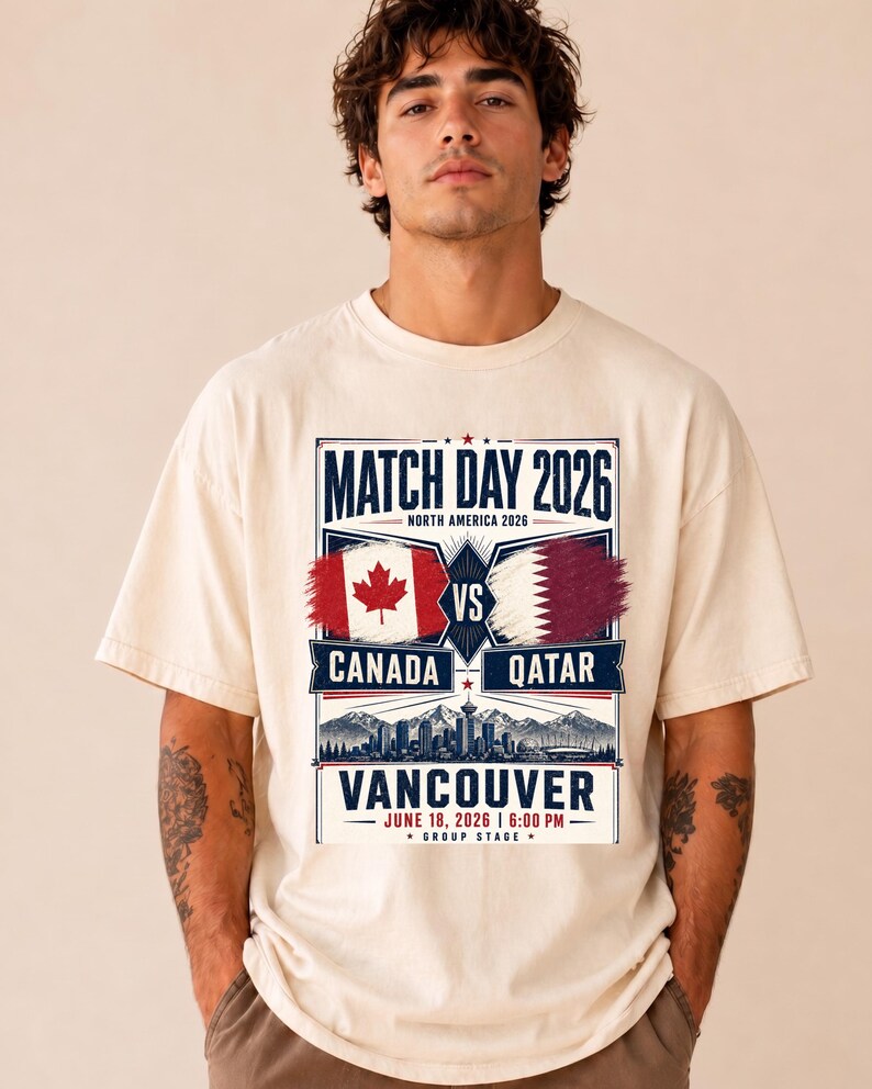 May include: Cream-colored t-shirt featuring a graphic for a 2026 soccer match between Canada and Qatar in Vancouver. The design includes flags, city skyline, and the text "Match Day 2026". The date and time of the match are also displayed.